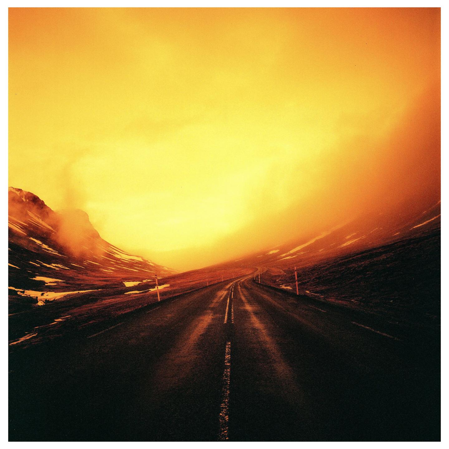 A road stretches into the distance through a mountainous landscape under a bright orange sky.