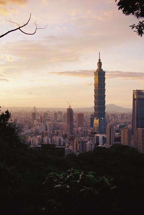 A tall skyscraper rises above a city skyline at sunset, viewed from a hillside with dark foliage in the foreground.