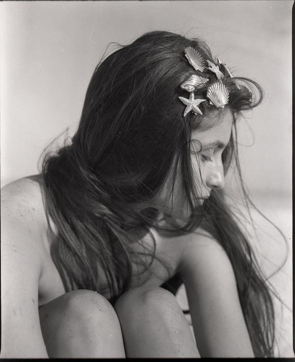 A woman with long hair and seashell hair clips sits with her knees drawn up.