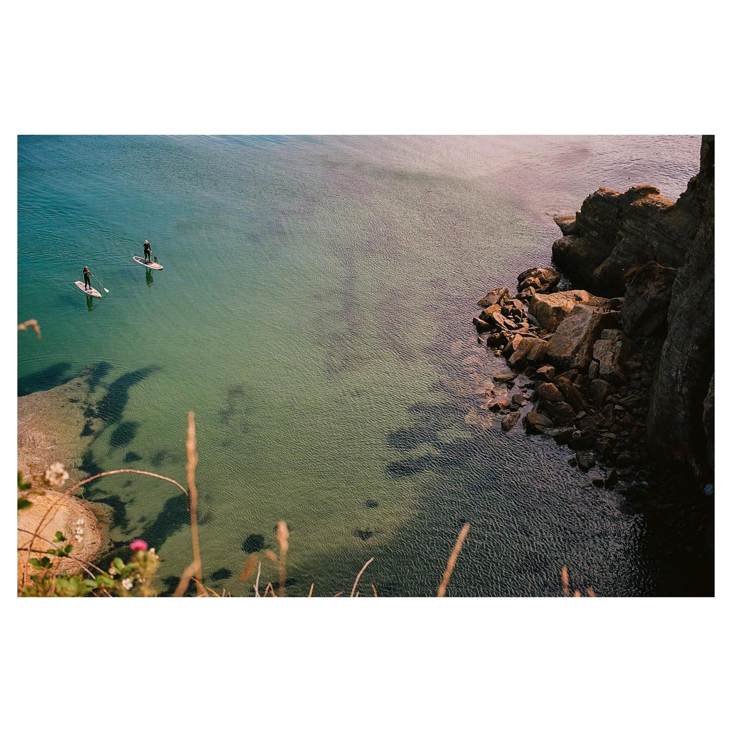 Two people paddleboard on clear water next to a rocky cliff.