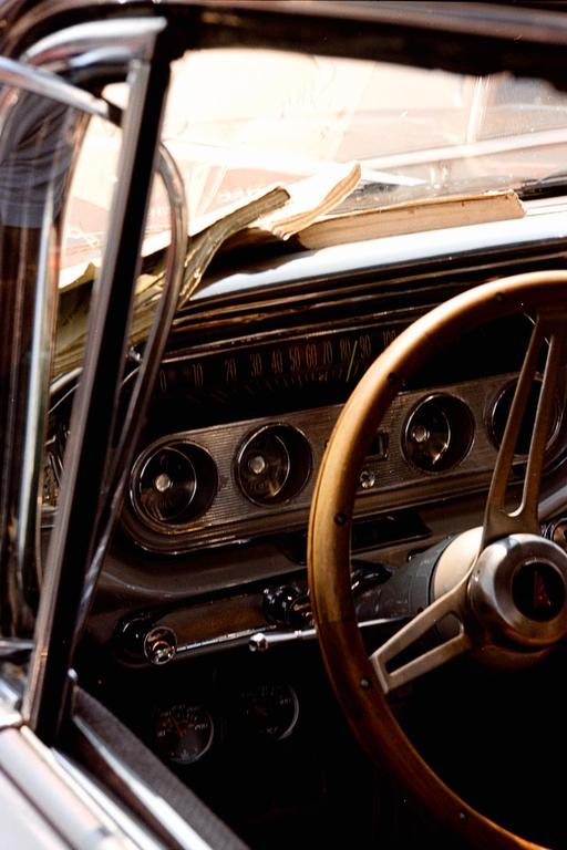 The interior of a vintage car shows the dashboard, steering wheel, and books on the windshield.