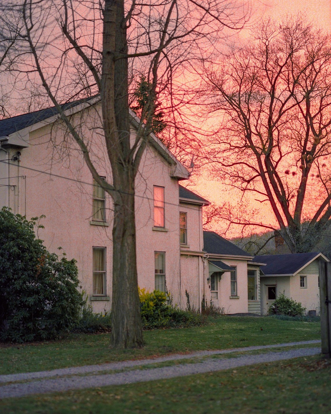 A house with a gravel driveway is surrounded by trees with a vibrant pink sky in the background.