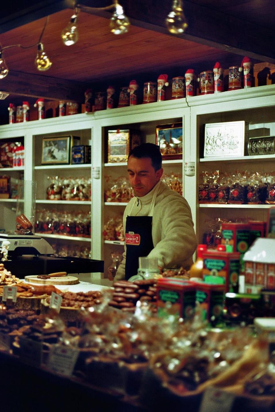 A man wearing an apron stands behind a counter filled with various baked goods and candies, with shelves of packaged items and Santa figurines in the background.