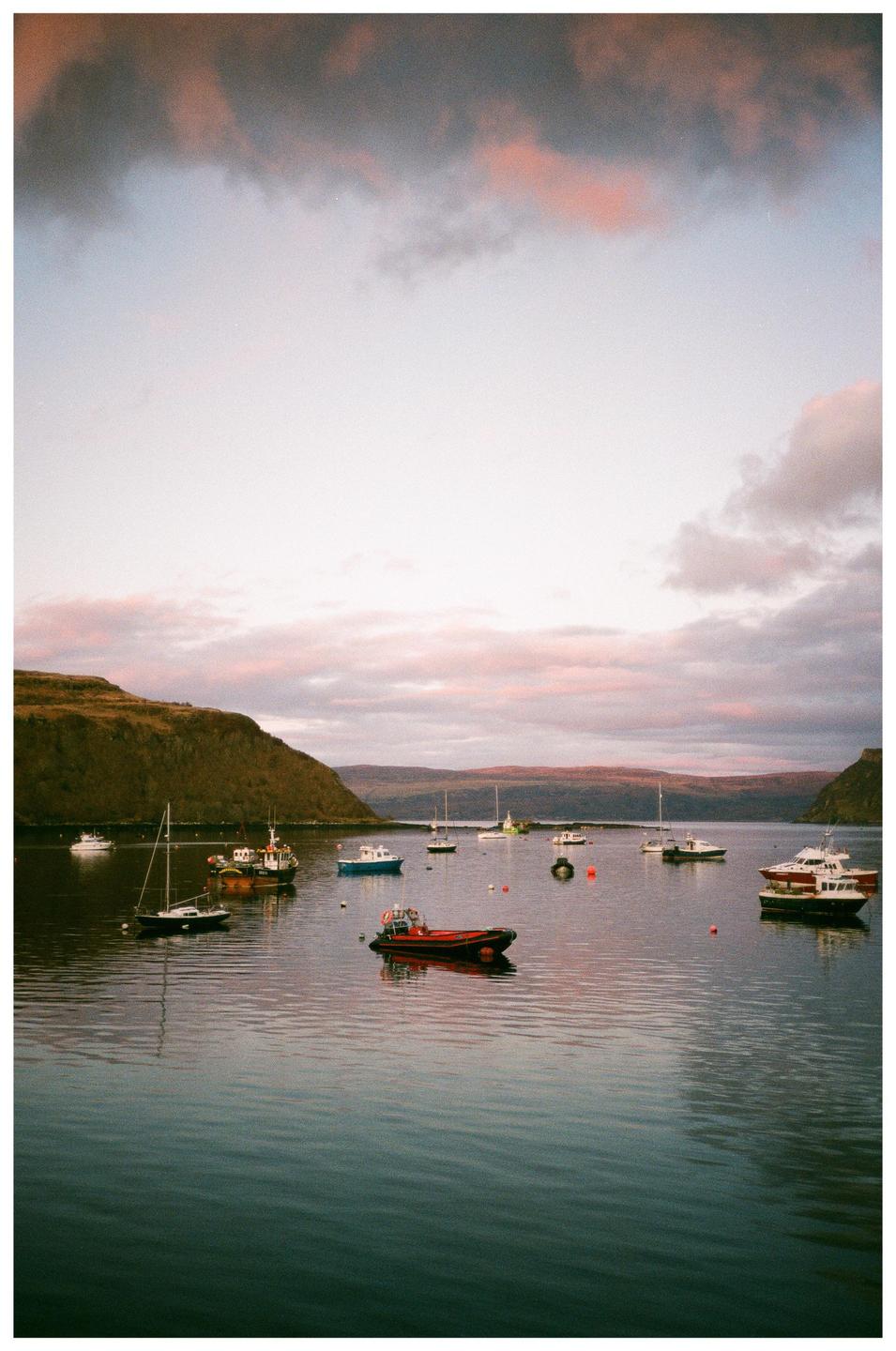 Boats are moored in a harbor with hills in the background under a cloudy sky.