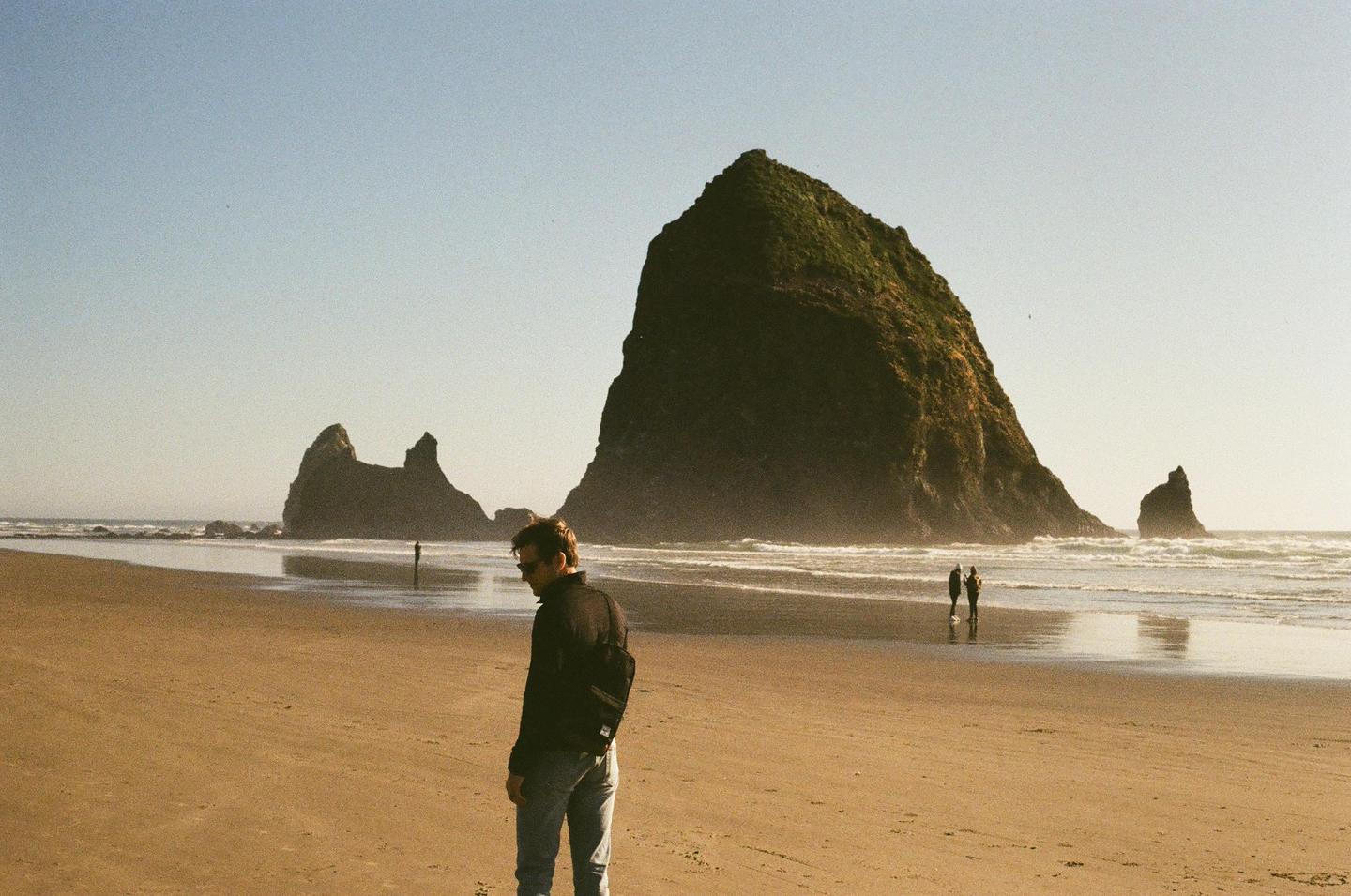 A man stands on a sandy beach with large rock formations in the background and the ocean to his right.