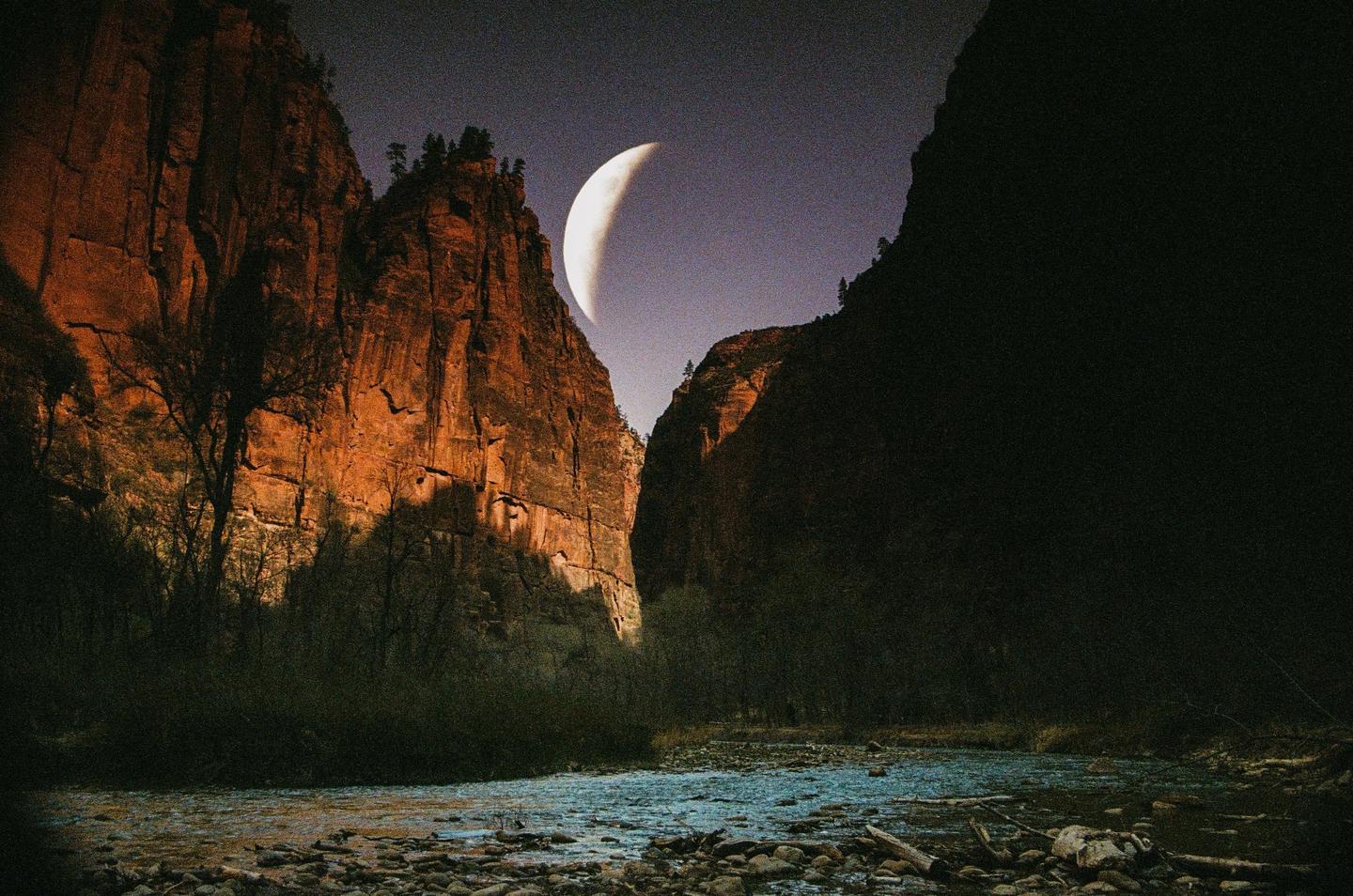 A crescent moon is visible in the night sky above a river flowing between two large rock formations.