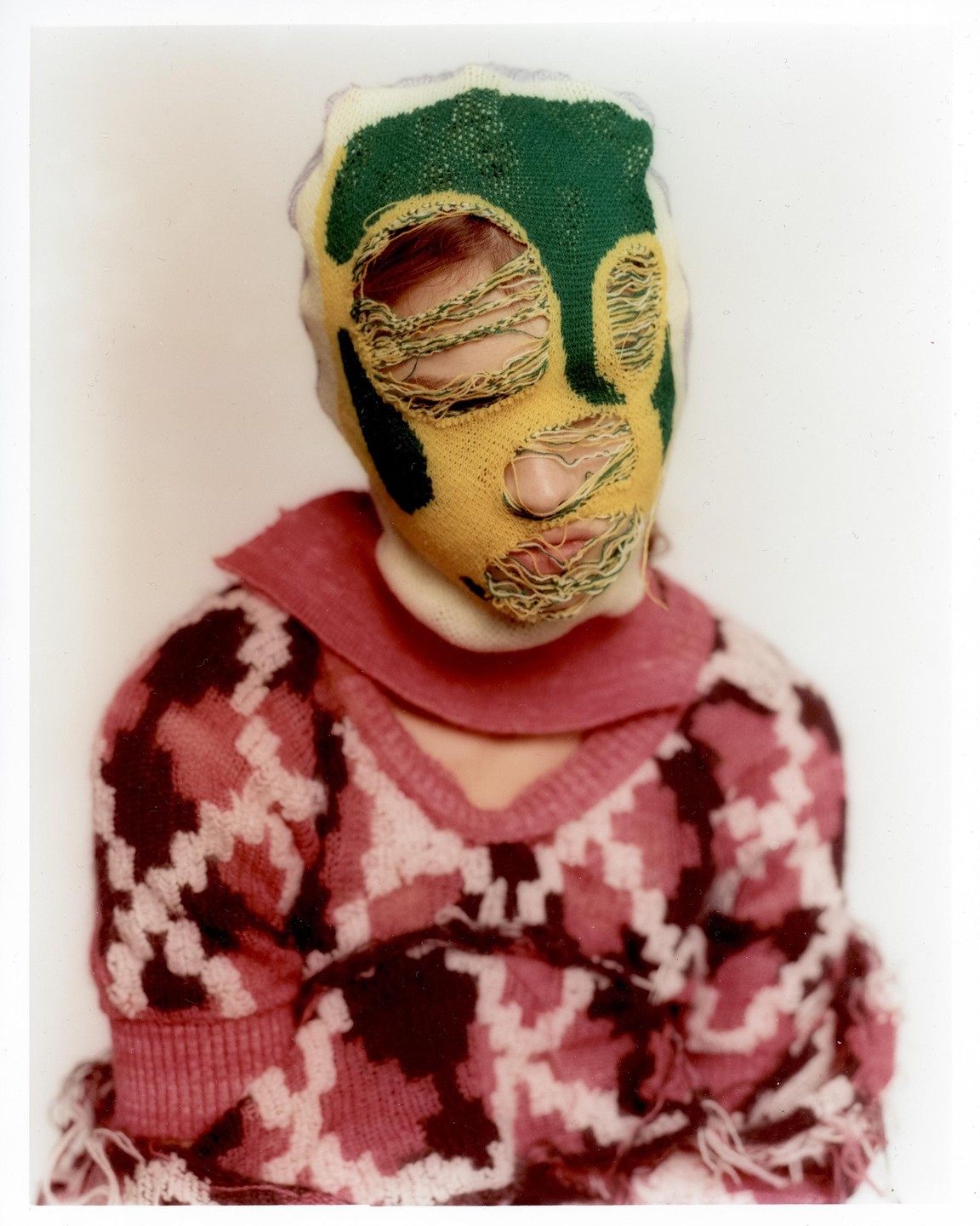 A person wears a green and yellow knitted mask with holes for eyes, nose, and mouth, paired with a pink and white patterned sweater.