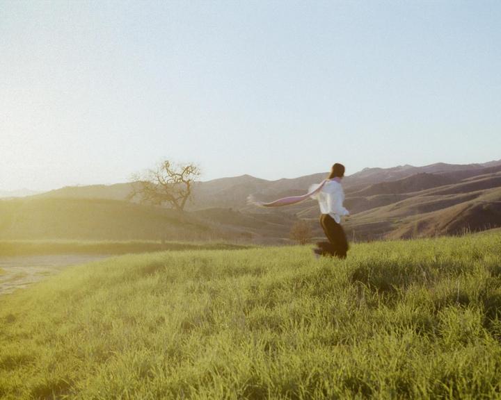 A woman jumps in a grassy field with rolling hills and a tree in the background.