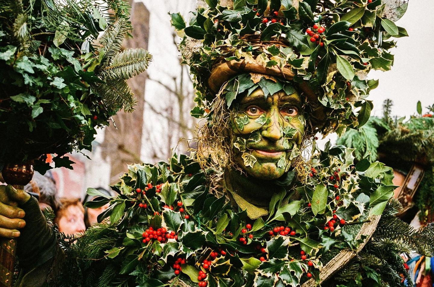 A man with green face paint and leaves on his face and body looks directly at the viewer.