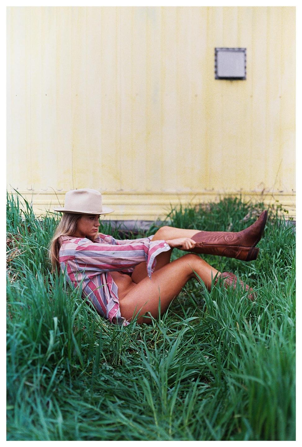 A woman wearing a cowboy hat, a striped shirt, and cowboy boots is lying in tall grass next to a yellow wall.