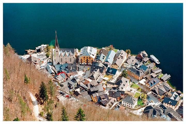 A village with a prominent church steeple is nestled on the shore of a large lake, surrounded by a forest of bare trees.