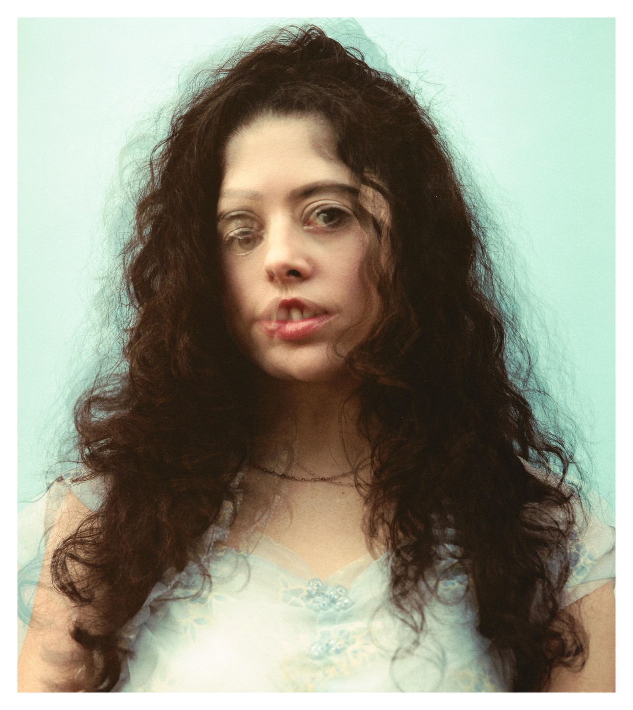 A woman with long, dark, curly hair wears a light blue dress and looks directly at the viewer with a double exposure effect.