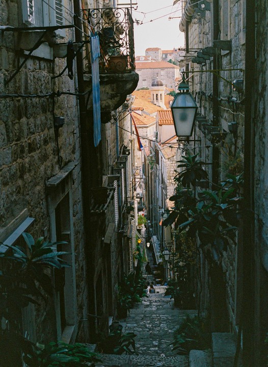 A narrow, cobbled street descends between stone buildings with terracotta roofs visible in the distance.