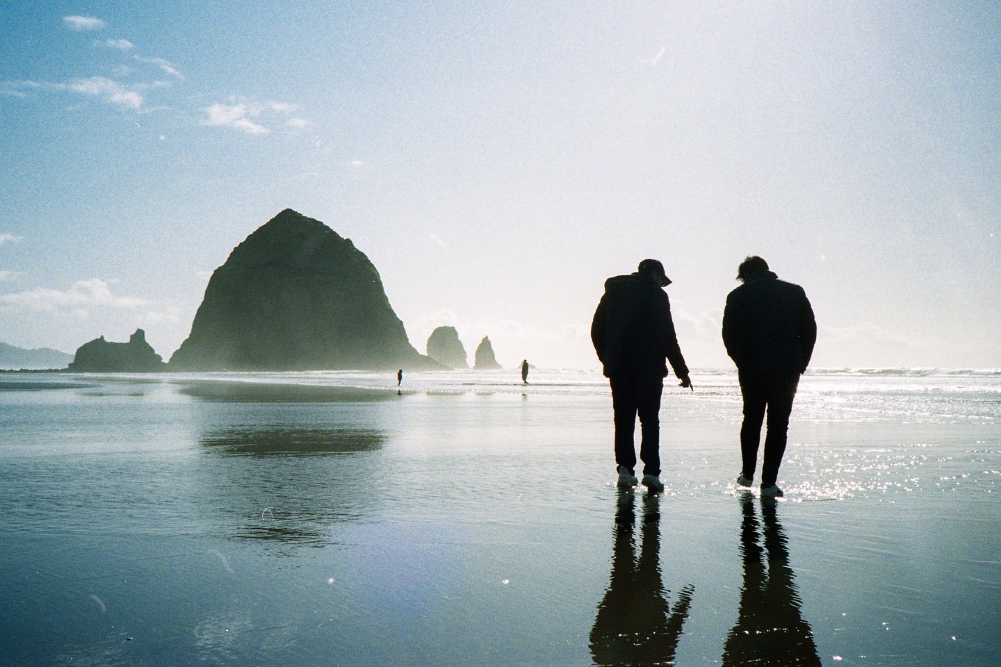 Two men walk on a wet beach with a large rock formation in the background.