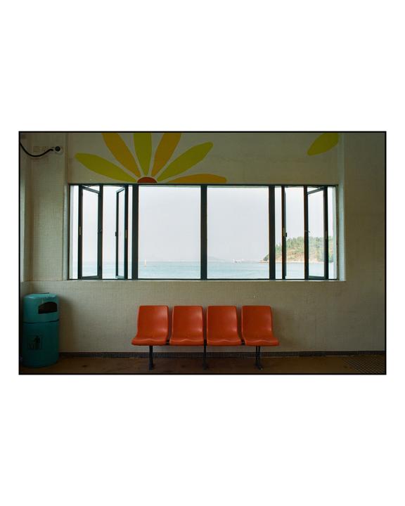 Four orange chairs sit in front of a window overlooking the ocean, with a yellow sun painted on the wall above the window.