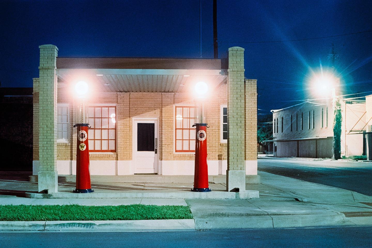 Two red vintage gas pumps with illuminated globes stand in front of a brick building at night.