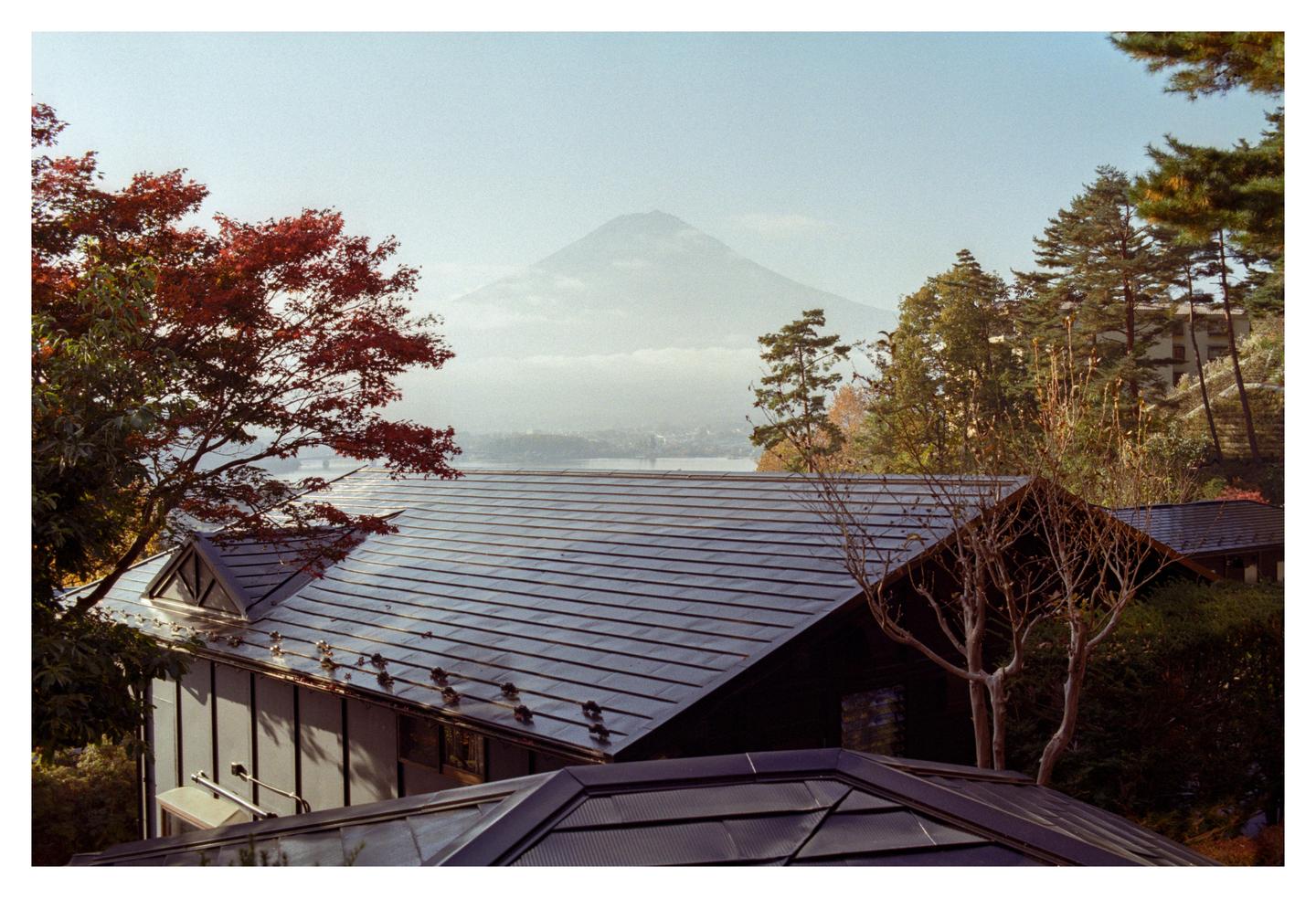 A mountain rises in the distance behind a lake and several buildings with dark roofs, surrounded by trees with autumn foliage.