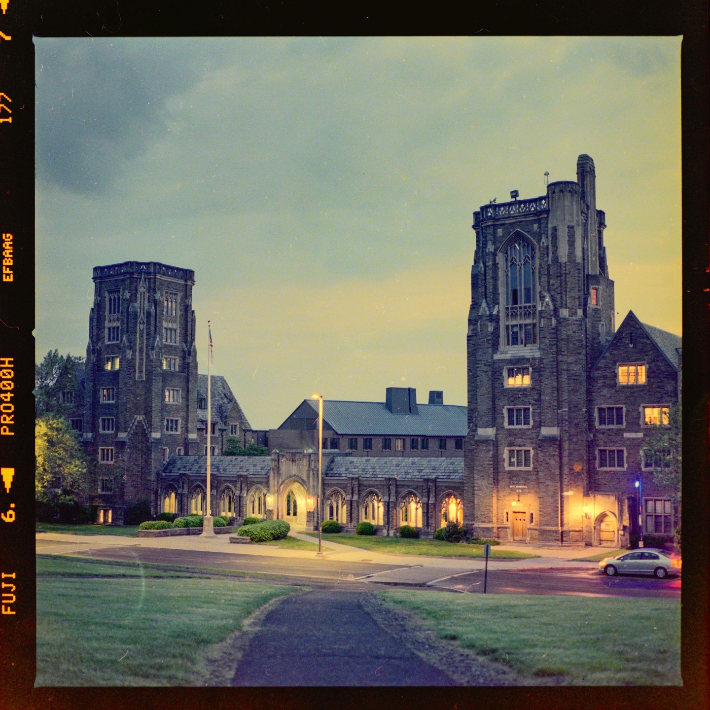 A large stone building with two prominent towers is illuminated at dusk, with a car parked on the right.
