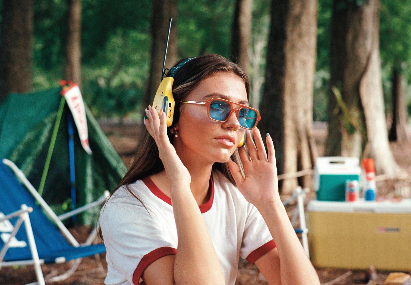 A woman wearing headphones and sunglasses sits outdoors with a tent and cooler in the background.