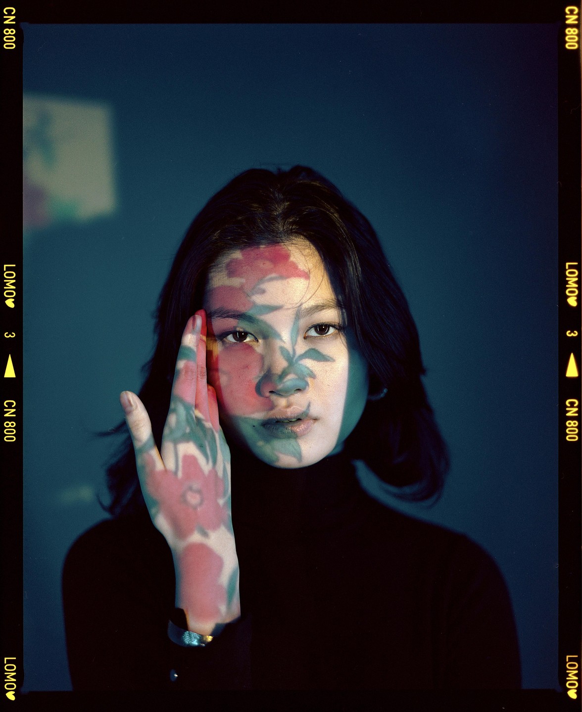 A woman with dark hair looks directly at the viewer, with a floral pattern projected onto her face and hand.