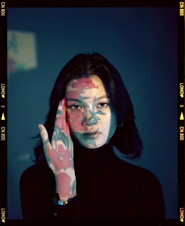 A woman with dark hair looks directly at the viewer, with a floral pattern projected onto her face and hand.