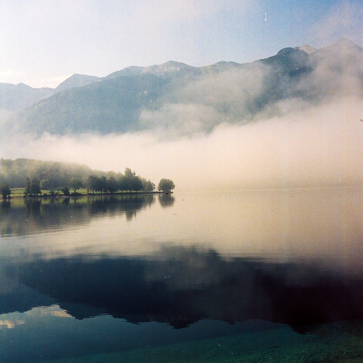 A foggy lake with mountains in the background and trees on the shore is reflected in the water.