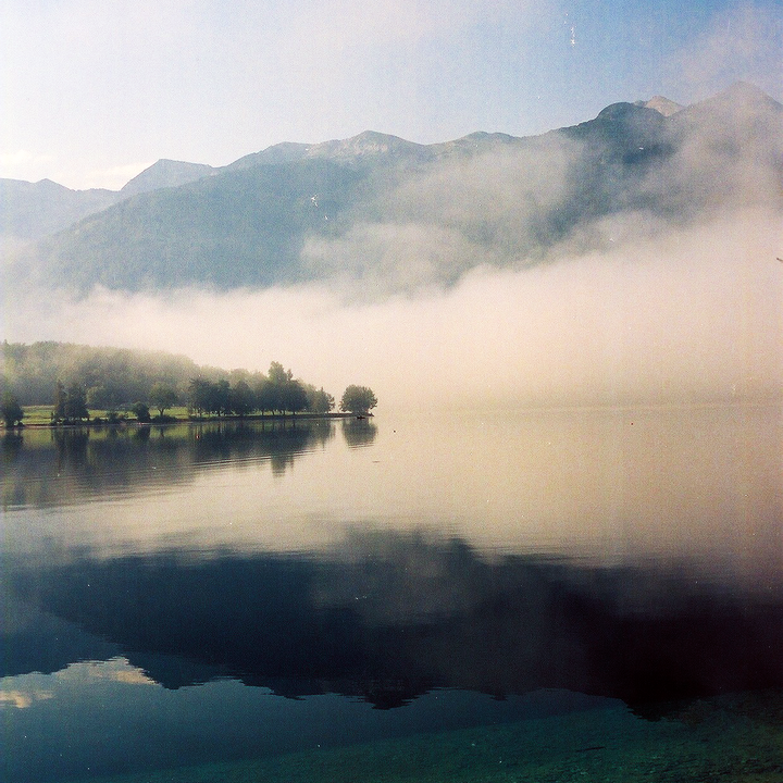 A foggy lake with mountains in the background and trees on the shore is reflected in the water.