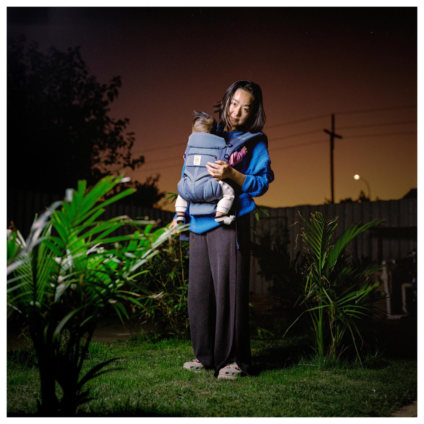 A woman holds a baby in a carrier in a garden at night.