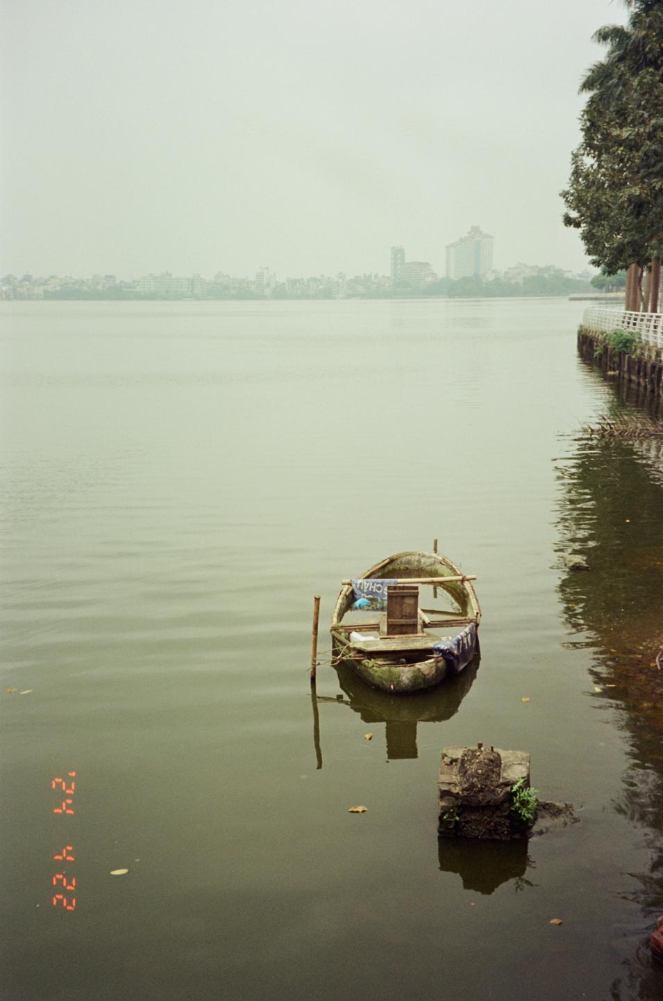 A small, old boat floats on a hazy lake with a city skyline in the distance.