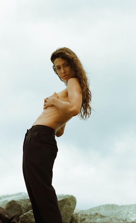 A woman with long, curly hair poses topless in black pants against a light blue sky.