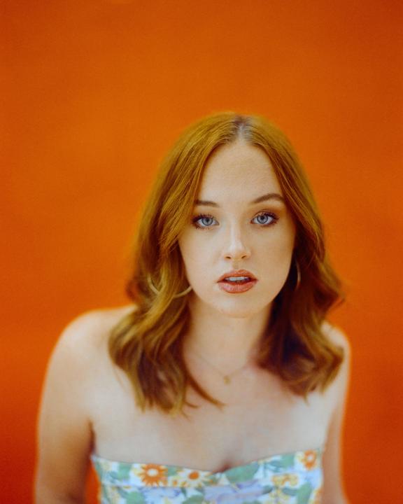 A woman with red hair and blue eyes looks directly at the viewer against an orange background.