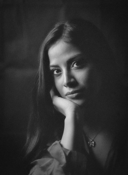 A woman with long hair rests her chin on her hand, looking directly at the viewer.