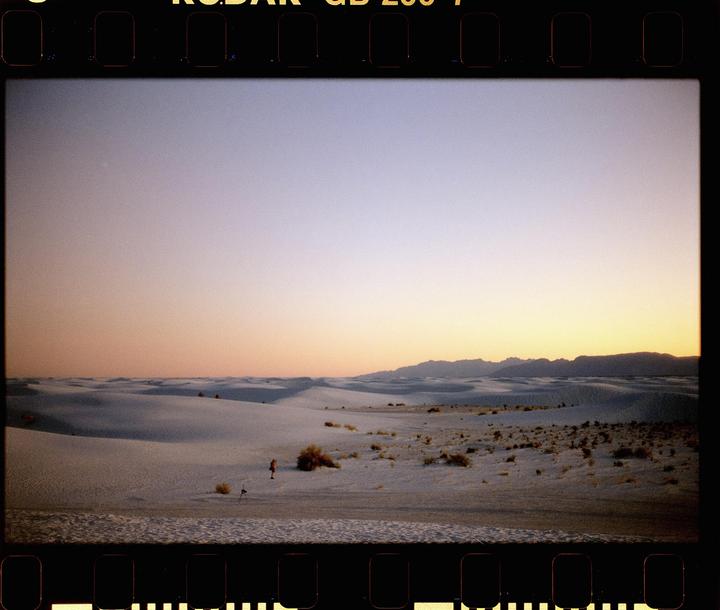A vast desert landscape with white sand dunes stretches towards mountains under a colorful sunset sky.