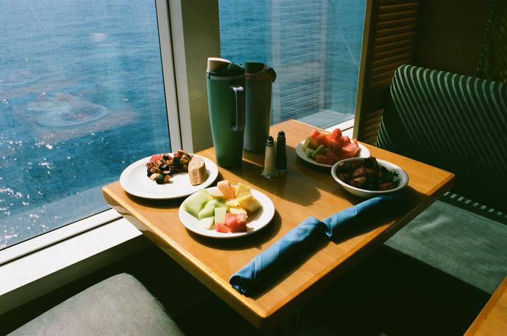 A table with plates of food and two thermoses sits next to a window overlooking the ocean.
