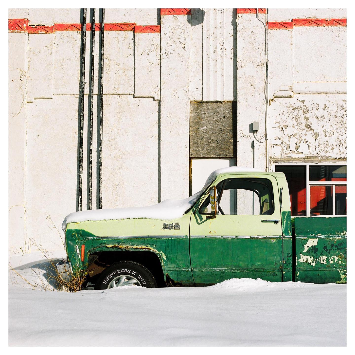 A snow-covered green and light green pickup truck is parked next to a white building with red trim.