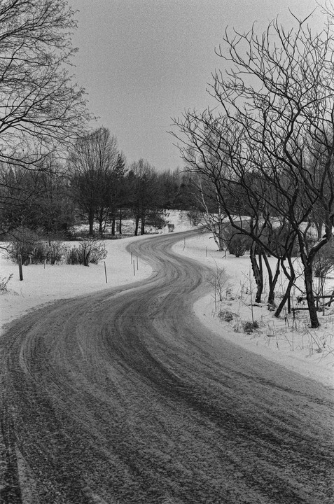 A winding road covered in snow leads through a winter landscape with bare trees.
