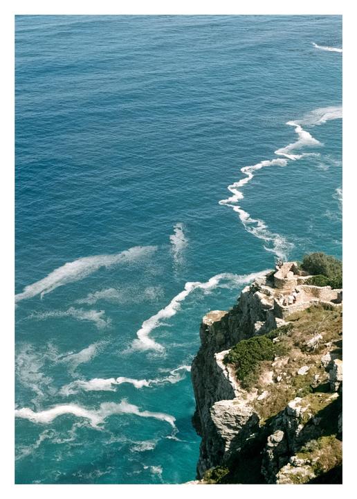 A high-angle view shows a rocky cliff overlooking a turquoise ocean with white foamy waves.