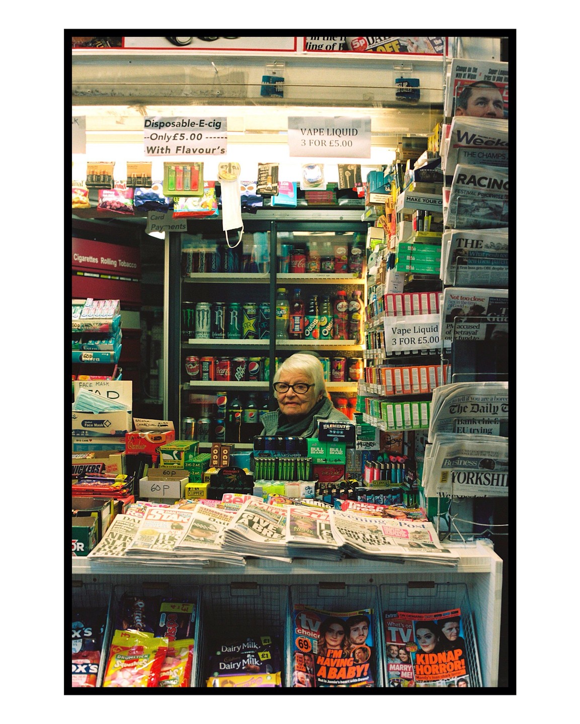 An elderly woman with glasses looks out from behind a counter filled with newspapers, magazines, and various convenience store items.