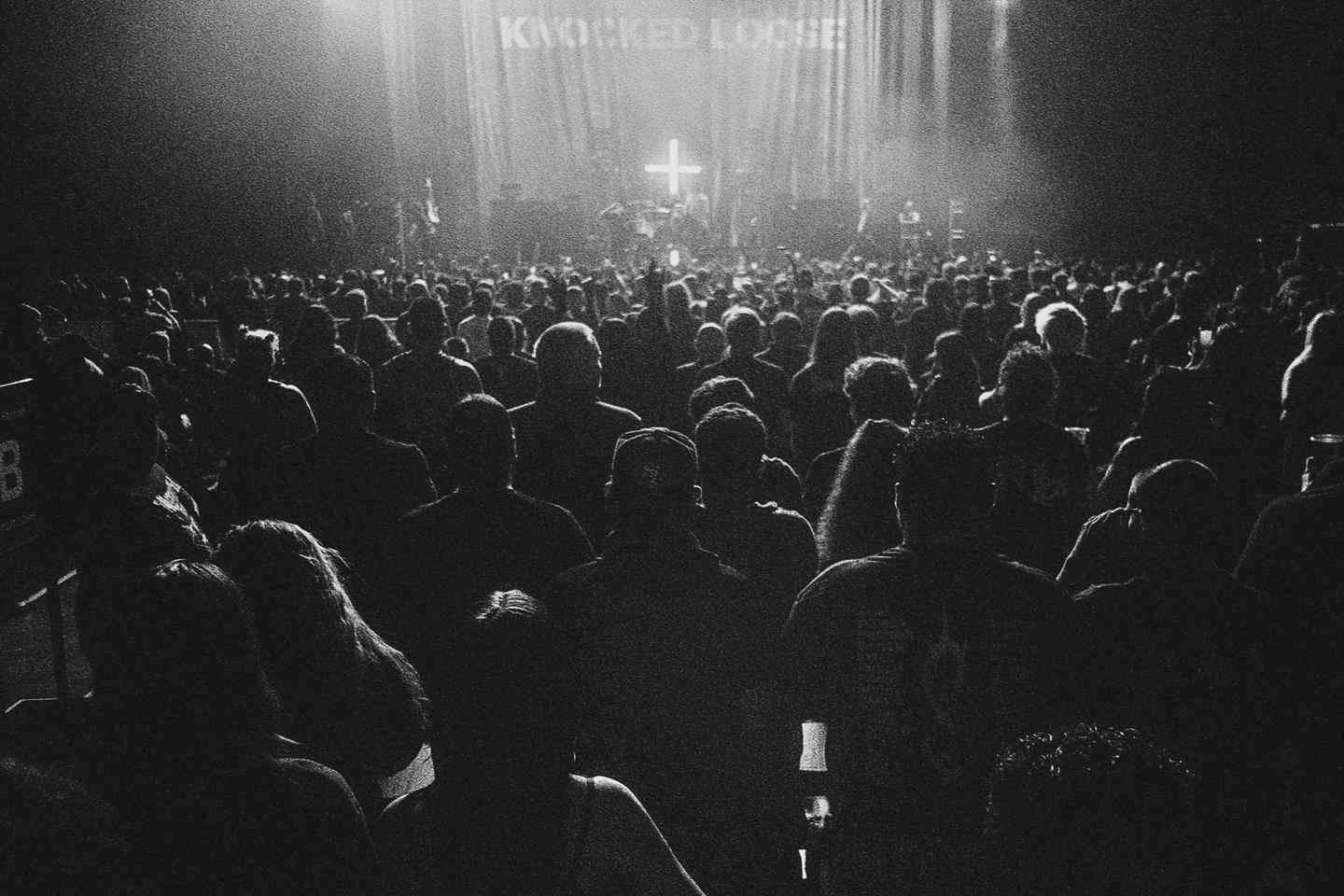 A large crowd of people stands in a dark venue, facing a brightly lit stage with a cross symbol and the words "KNOCKED LOOSE" visible.