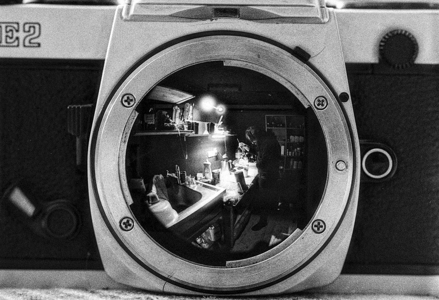 A person works in a darkroom, reflected in the lens mount of a camera.