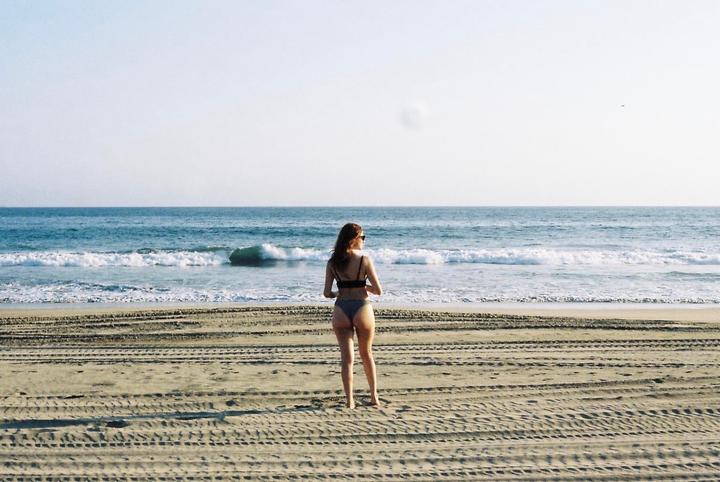 A woman in a bikini stands on a sandy beach looking out at the ocean waves.