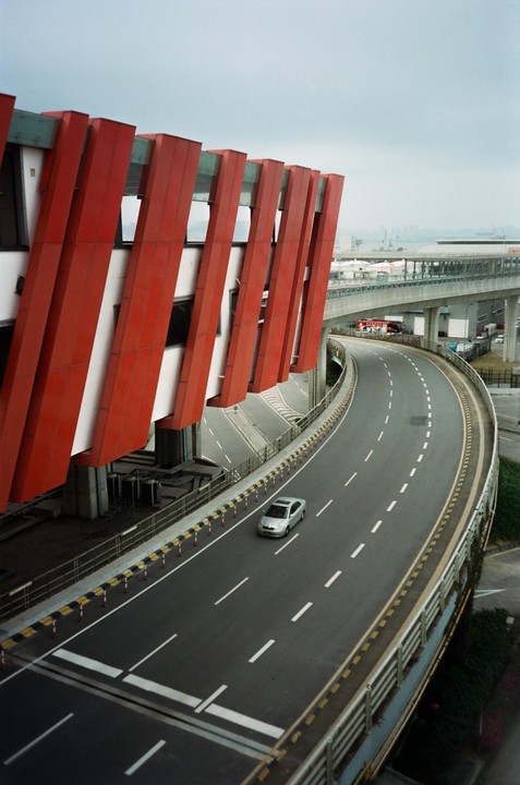 A silver car drives on a curved road next to a building with red architectural elements.
