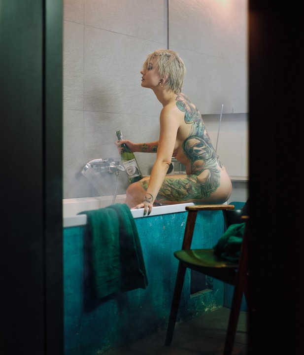 A woman with tattoos sits on the edge of a bathtub, holding a bottle of wine.