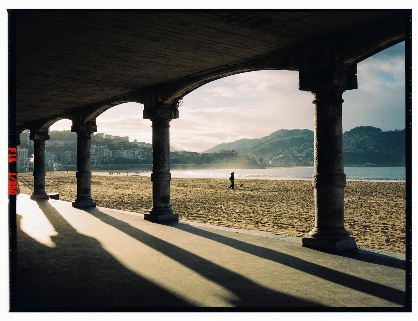 A person walks a dog on a sandy beach with mountains and a city in the background, viewed from under an arched promenade.