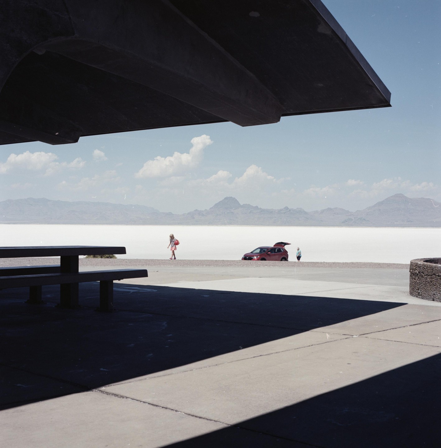 A woman walks past a red car with its trunk open on a salt flat with mountains in the background, viewed from under a concrete overhang.