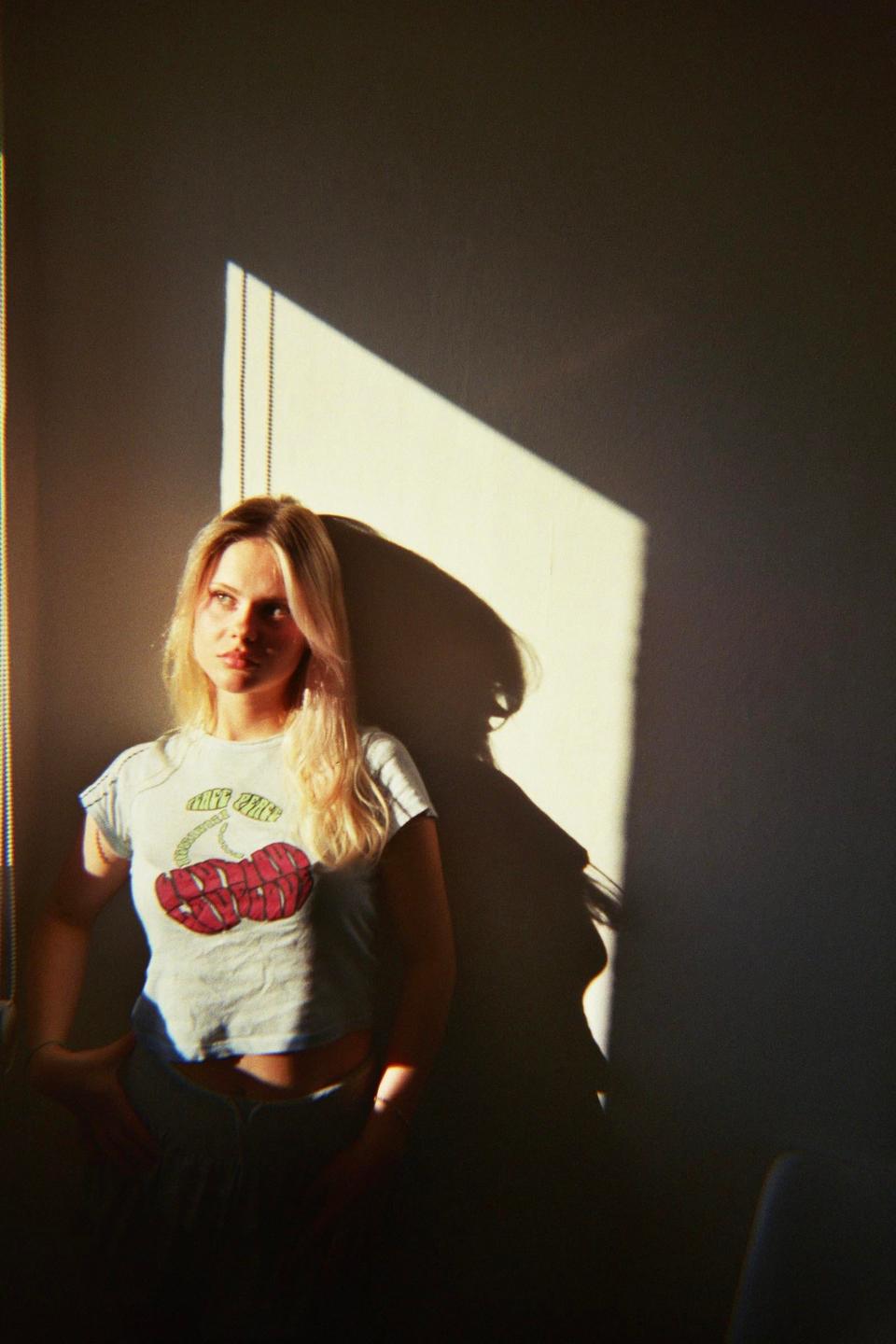 A woman with long blonde hair stands against a wall with a strong shadow cast upon it.