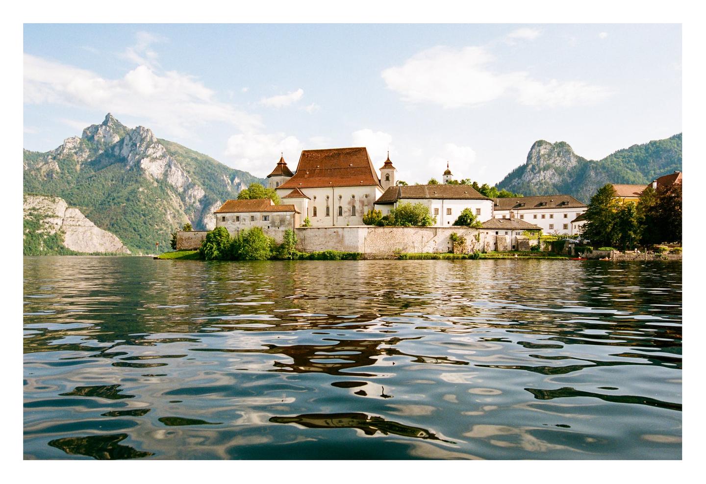 A large building with a red roof sits on the edge of a lake, with mountains in the background.