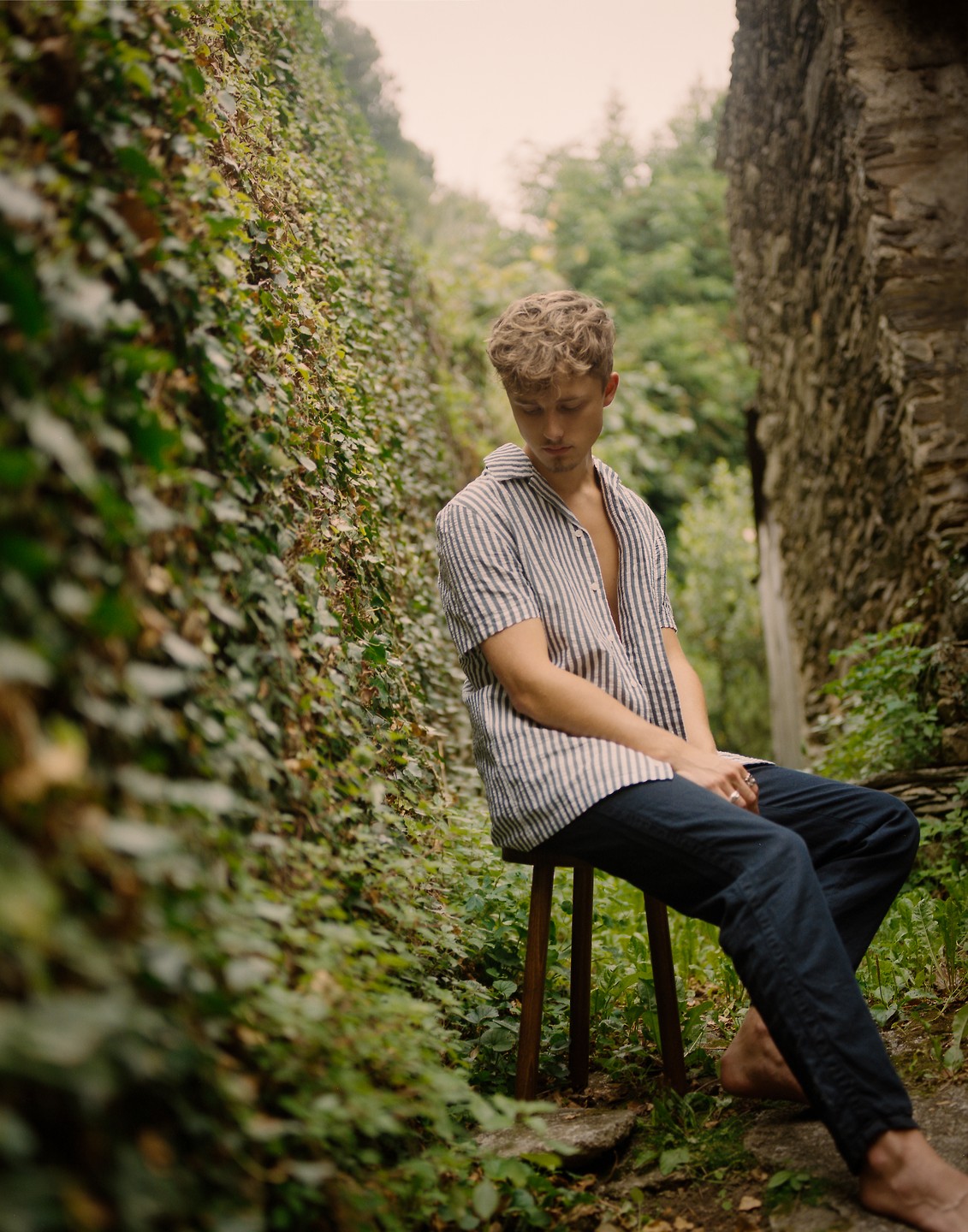 A man with curly hair sits on a stool in a narrow, overgrown alleyway with stone walls.