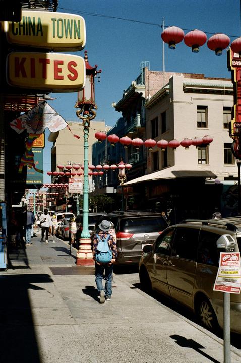 A person with a blue backpack walks down a street lined with parked cars and buildings adorned with red lanterns.