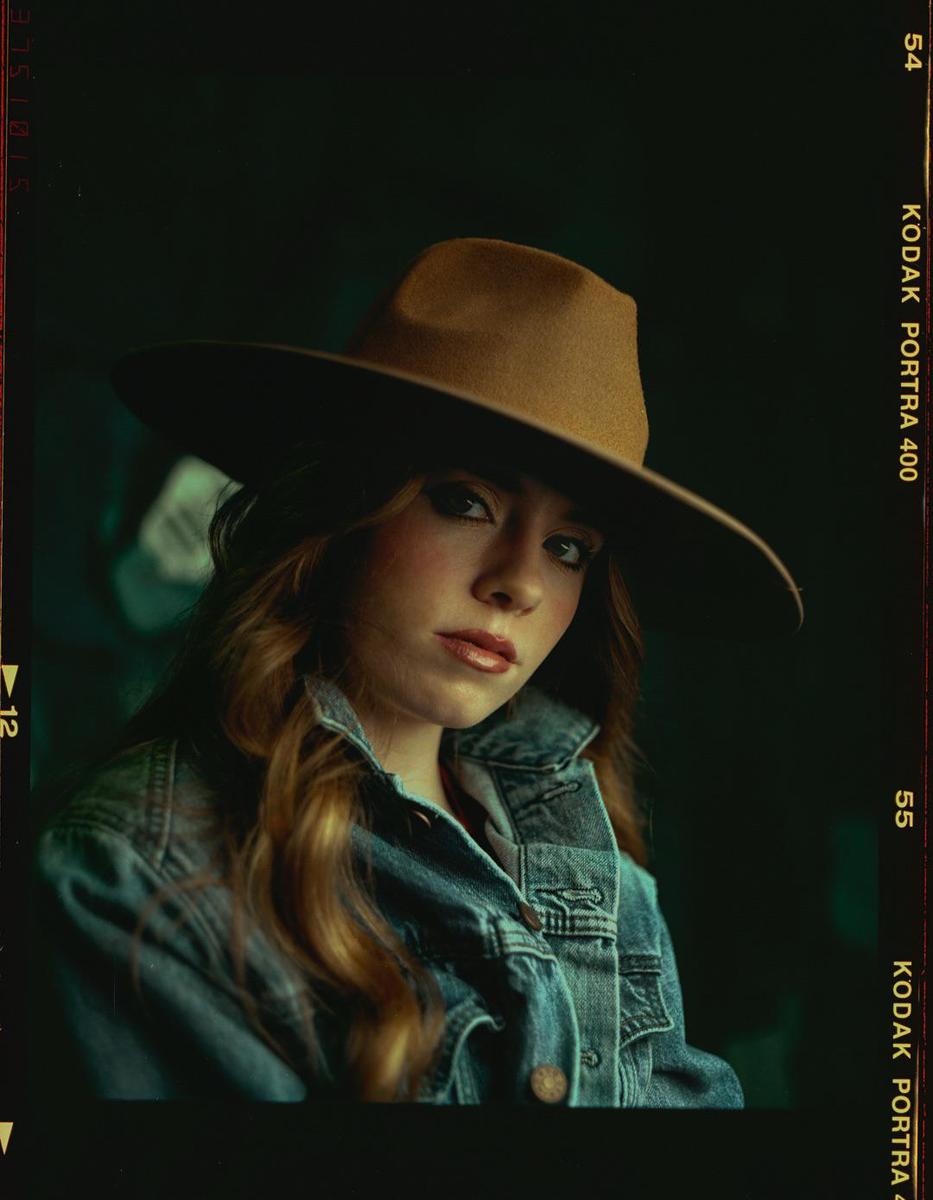 A woman with long hair and a denim jacket wears a wide-brimmed hat.